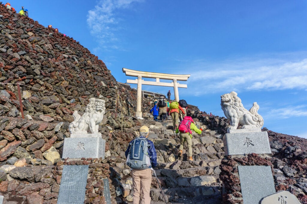 Des randonneurs gravissent un chemin de pierres menant à une porte torii blanche, flanquée de deux statues de chiens-lions sculptés, sous un ciel bleu. Située dans les Alpes japonaises, la scène évoque des sommets sacrés avec d'autres personnes visibles à l'arrière-plan. - Karavaniers