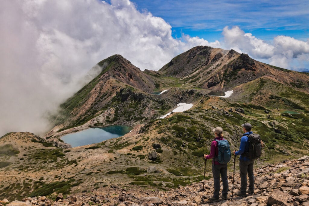 Deux randonneurs se tiennent sur une pente rocheuse dans les Alpes japonaises, contemplant un paysage accidenté composé de terrains verts et bruns, de taches de neige, d'un petit lac bleu et d'un ciel partiellement nuageux - une vue impressionnante parmi les sommets sacrés. - Karavaniers
