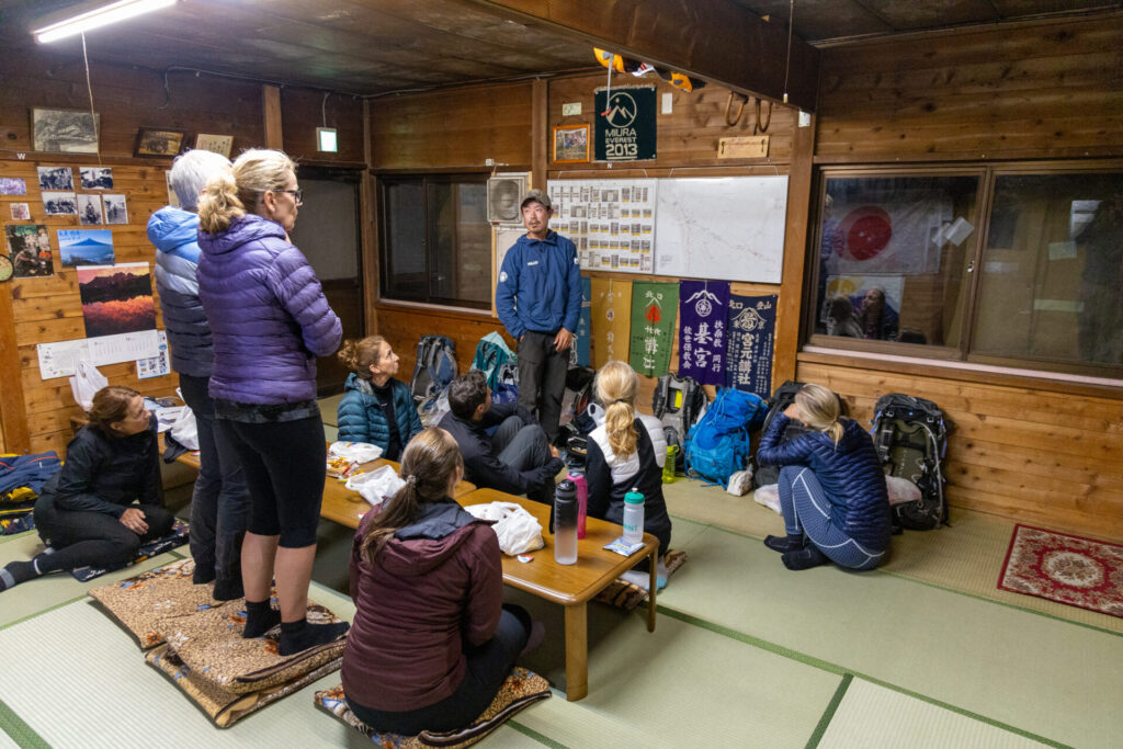 Dans une salle en bois, un groupe de personnes en tenue de plein air est assis et debout autour de tables basses, écoutant une personne parler près d'un mur couvert de cartes, d'affiches et de matériel de randonnée des Alpes japonaises et de leurs sommets sacrés. - Karavaniers