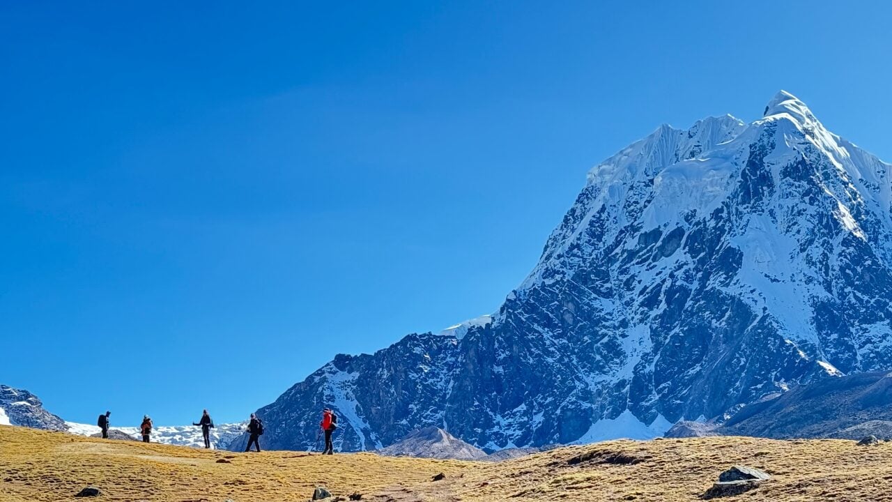 Cinq personnes se promènent dans une plaine herbeuse avec, en arrière-plan, un pic montagneux enneigé et un ciel bleu limpide - une scène incontournable pour tout amoureux de la nature après une conférence ou une retraite. - Karavaniers