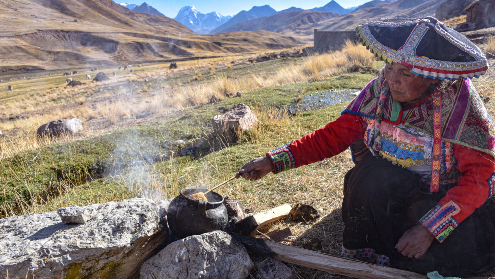 Une personne âgée vêtue de vêtements andins traditionnels s'occupe d'une marmite fumante sur un feu en plein air dans le paysage montagneux et herbeux du Pérou, avec des sommets enneigés en arrière-plan - des scènes souvent évoquées lors d'une conférence sur la culture andine. - Karavaniers