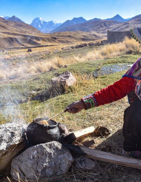 Une personne âgée vêtue de vêtements andins traditionnels s'occupe d'une marmite fumante sur un feu en plein air dans le paysage montagneux et herbeux du Pérou, avec des sommets enneigés en arrière-plan - des scènes souvent évoquées lors d'une conférence sur la culture andine. - Karavaniers