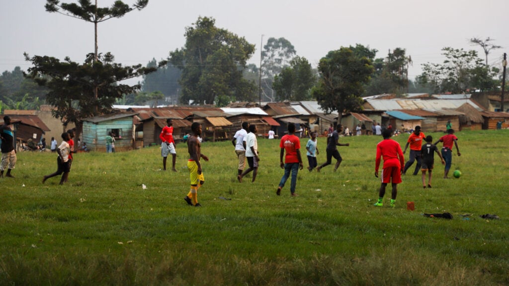 Un groupe d'enfants et d'adolescents jouent au football sur un terrain herbeux avec des maisons et des arbres en arrière-plan. Certains joueurs portent des vêtements rouges et une personne en jaune se tient près du ballon. La scène semble animée et décontractée. - Karavaniers