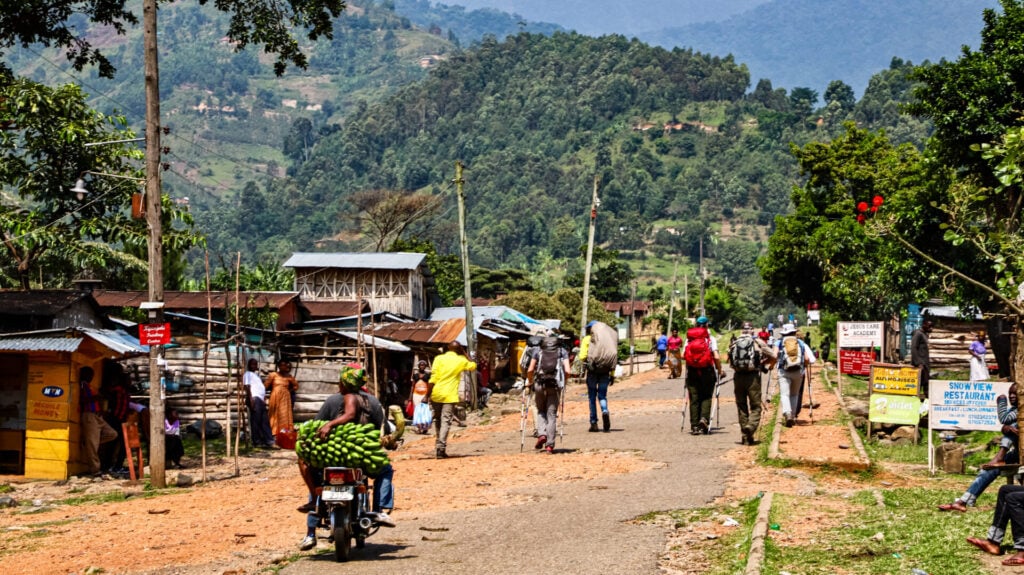 Une route rurale bordée de petits bâtiments en bois, des gens marchant avec des sacs à dos, un homme à moto transportant des bananes vertes, et des collines vertes en arrière-plan sous un ciel partiellement nuageux. - Karavaniers