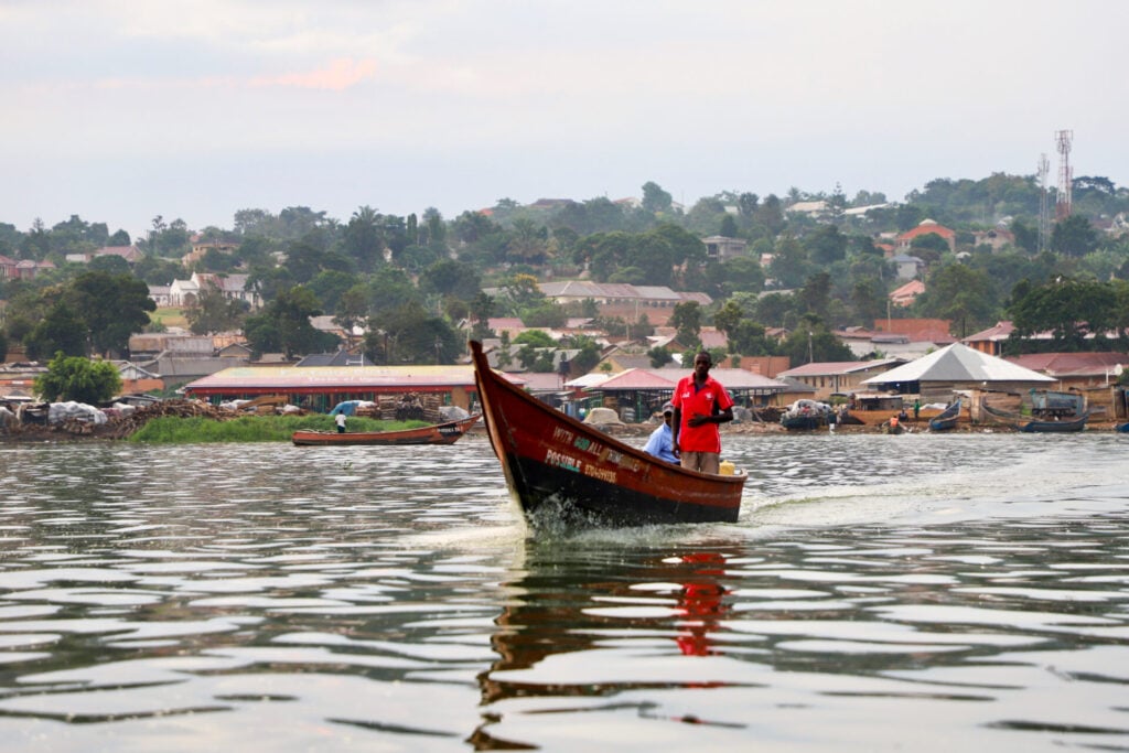 Un bateau en bois avec deux personnes se déplace sur un plan d'eau calme. À l'arrière-plan, des maisons, des arbres et des bateaux longent le rivage sous un ciel nuageux. - Karavaniers