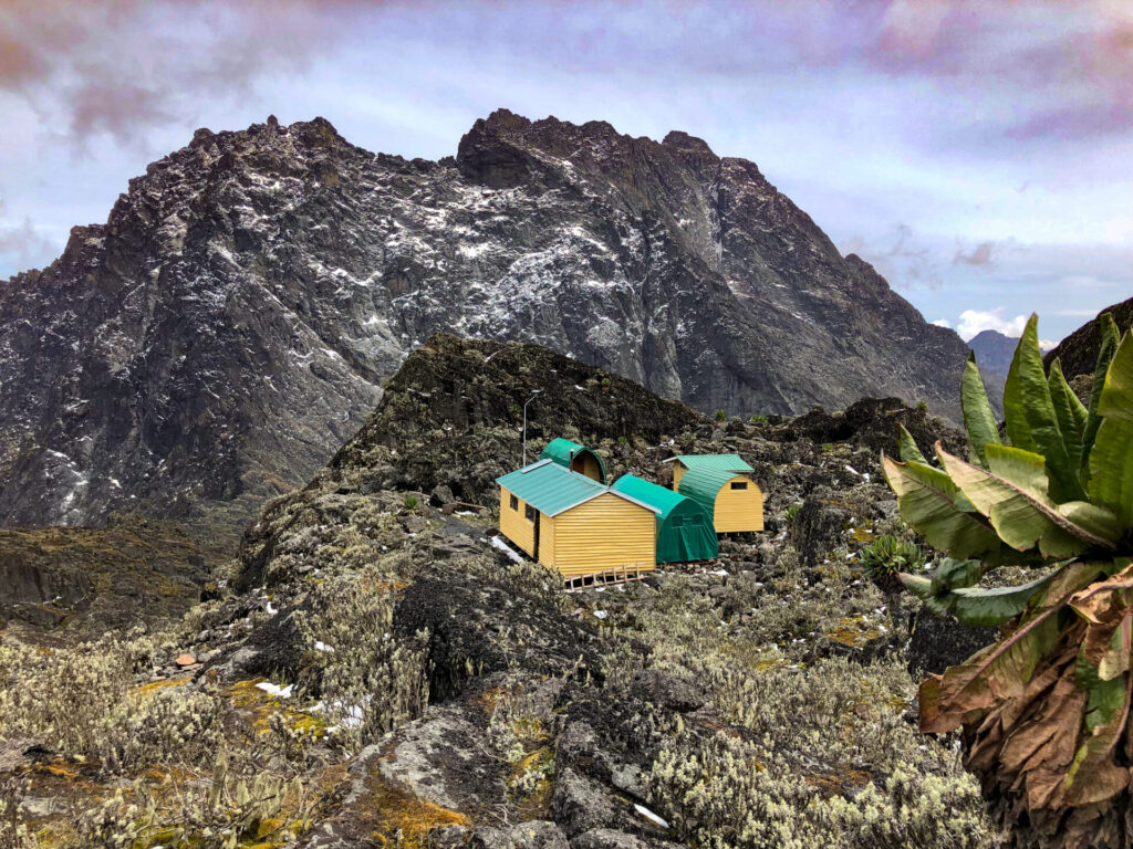 Plusieurs petites cabanes jaunes aux toits verts se dressent sur un terrain rocheux et montagneux au milieu des montagnes. De grands sommets escarpés s'élèvent à l'arrière-plan, tandis qu'une végétation clairsemée est visible au premier plan. Le ciel est partiellement nuageux. - Karavaniers