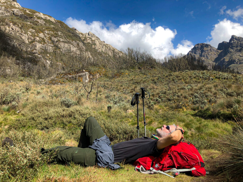 Une personne est allongée sur l'herbe, les mains derrière la tête, à côté d'un sac à dos rouge et de bâtons de randonnée, entourée de montagnes et d'un ciel bleu nuageux. - Karavaniers