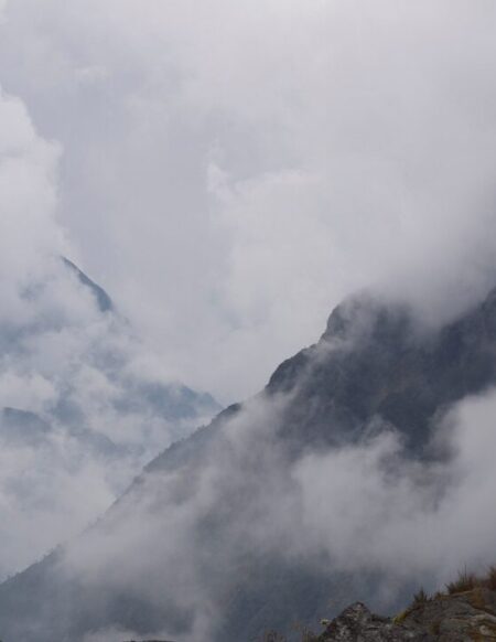 Des nuages cachent partiellement des pics montagneux rocheux et déchiquetés. Le ciel est couvert et le premier plan comprend des rochers sombres et une végétation clairsemée, rappelant les remarquables oubliés explorés lors de notre dernière conférence. - Karavaniers