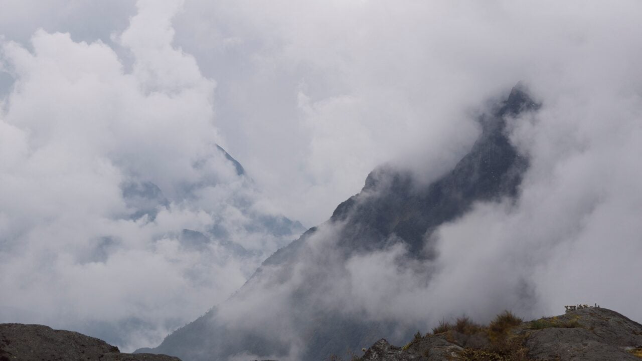 Des nuages cachent partiellement des pics montagneux rocheux et déchiquetés. Le ciel est couvert et le premier plan comprend des rochers sombres et une végétation clairsemée, rappelant les remarquables oubliés explorés lors de notre dernière conférence. - Karavaniers