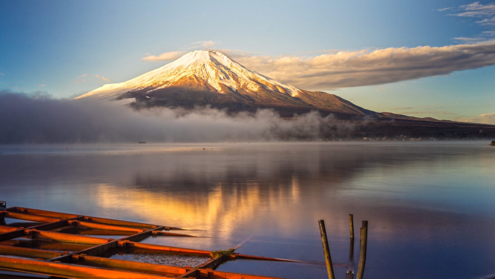 Le Mont Fuji enneigé se reflète dans un lac calme au lever du soleil, avec de la brume sur l'eau et une structure en bois orange au premier plan, capturant la beauté sereine du Japon. - Karavaniers