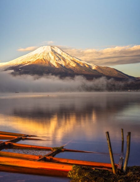 Le Mont Fuji enneigé se reflète dans un lac calme au lever du soleil, avec de la brume sur l'eau et une structure en bois orange au premier plan, capturant la beauté sereine du Japon. - Karavaniers