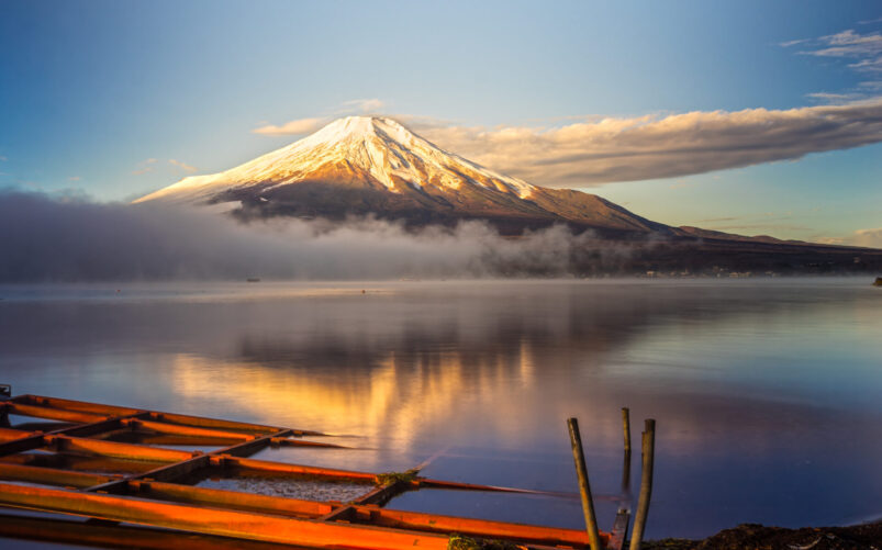 Le Mont Fuji enneigé se reflète dans un lac calme au lever du soleil, avec de la brume sur l'eau et une structure en bois orange au premier plan, capturant la beauté sereine du Japon. - Karavaniers