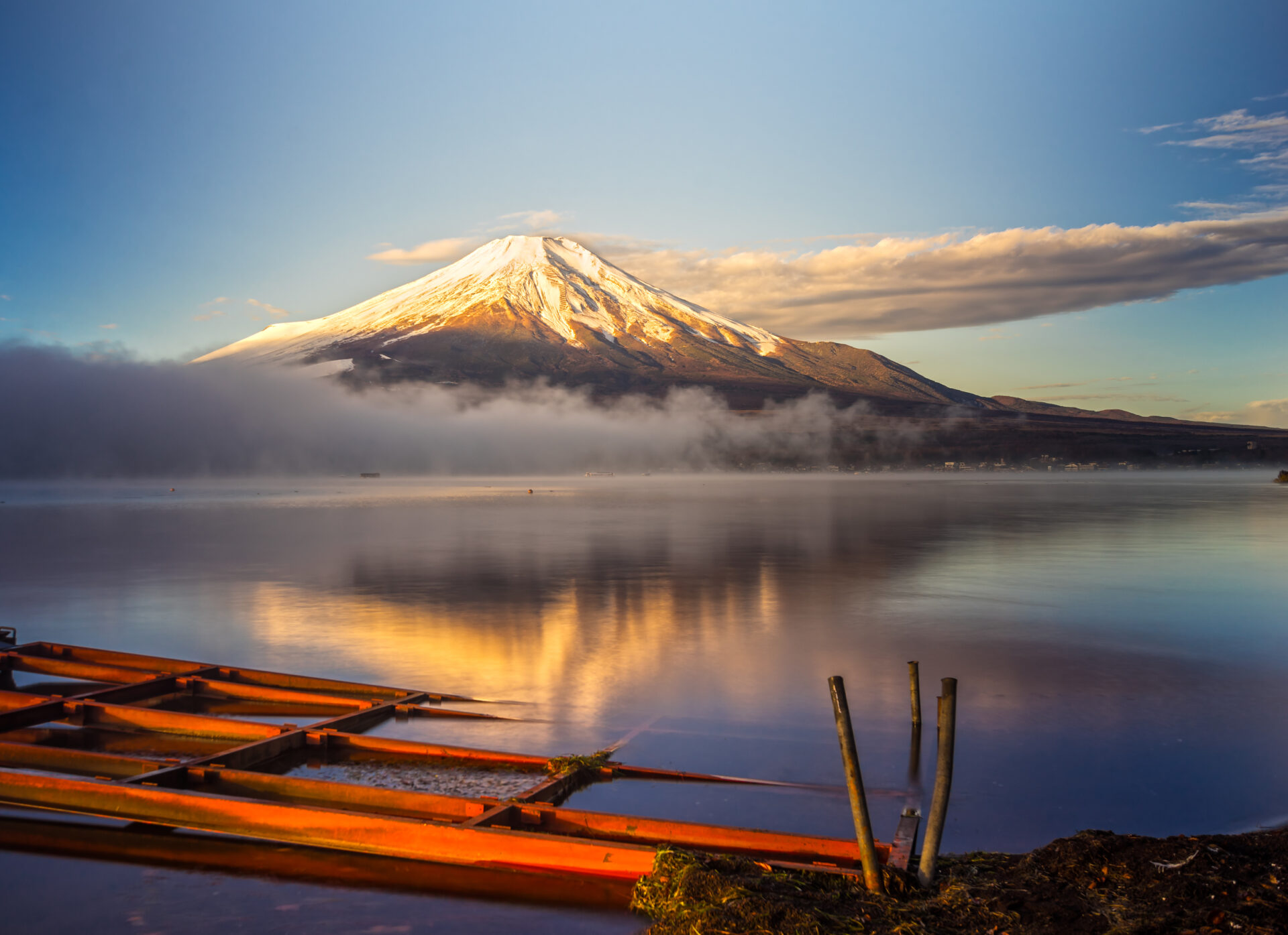 Le Mont Fuji enneigé se reflète dans un lac calme au lever du soleil, avec de la brume sur l'eau et une structure en bois orange au premier plan, capturant la beauté sereine du Japon. - Karavaniers