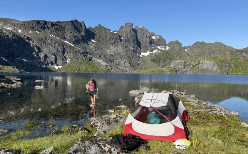 Une personne se tient au bord d'un lac de montagne aux eaux limpides, près d'une tente rouge et blanche dressée sur un terrain herbeux, avec du matériel de camping à proximité — une image qui évoque l'esprit d'un « Voyage au Maroc en famille » sous des falaises rocheuses, des plaques de neige et un ciel d'un bleu éclatant. - Karavaniers
