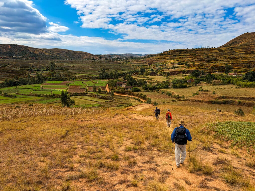 Quatre personnes descendent un sentier de terre au milieu des herbes sèches, s'embarquant dans un voyage mystérieux qui rappelle Madagascar, en direction des champs et des collines lointaines, sous un ciel partiellement nuageux. - Karavaniers