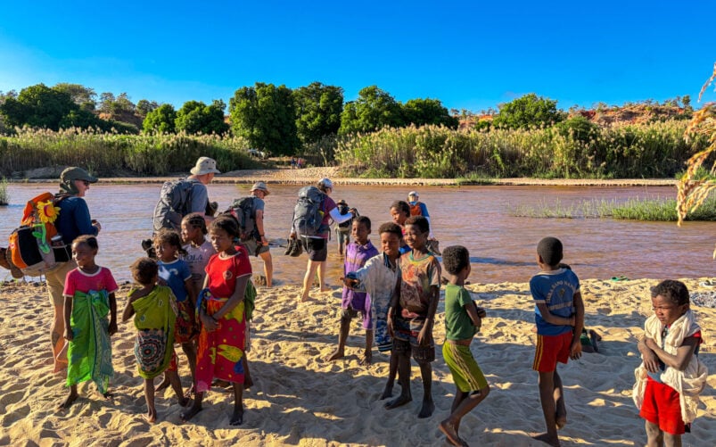 Un groupe d'enfants est debout et joue sur une berge sablonneuse, tandis que plusieurs adultes munis de sacs à dos — peut-être en route pour un voyage mystère à Madagascar — marchent à proximité ; une végétation luxuriante et une rivière boueuse se dessinent en arrière-plan, sous un ciel bleu limpide. - Karavaniers