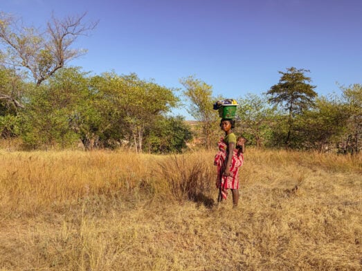 Une femme vêtue d'une robe rouge à motifs se tient debout dans un champ d'herbe sèche, un panier vert sur la tête et un bébé sur le dos : une scène tirée du « voyage mystère » à travers Madagascar, avec des arbres et des arbustes sous un ciel bleu limpide. - Karavaniers