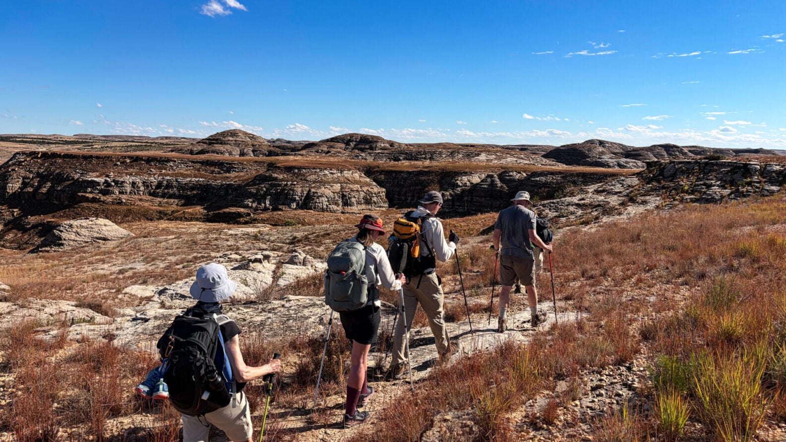 Quatre randonneurs, équipés de sacs à dos et de chapeaux, se lancent dans un voyage mystère sur un sentier rocailleux et herbeux, sous un ciel bleu limpide, en direction des collines lointaines qui se profilent dans le paysage aride et dégagé de Madagascar. - Karavaniers