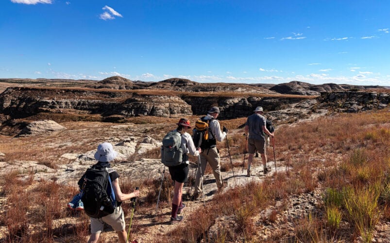 Quatre randonneurs, équipés de sacs à dos et de chapeaux, se lancent dans un voyage mystère sur un sentier rocailleux et herbeux, sous un ciel bleu limpide, en direction des collines lointaines qui se profilent dans le paysage aride et dégagé de Madagascar. - Karavaniers