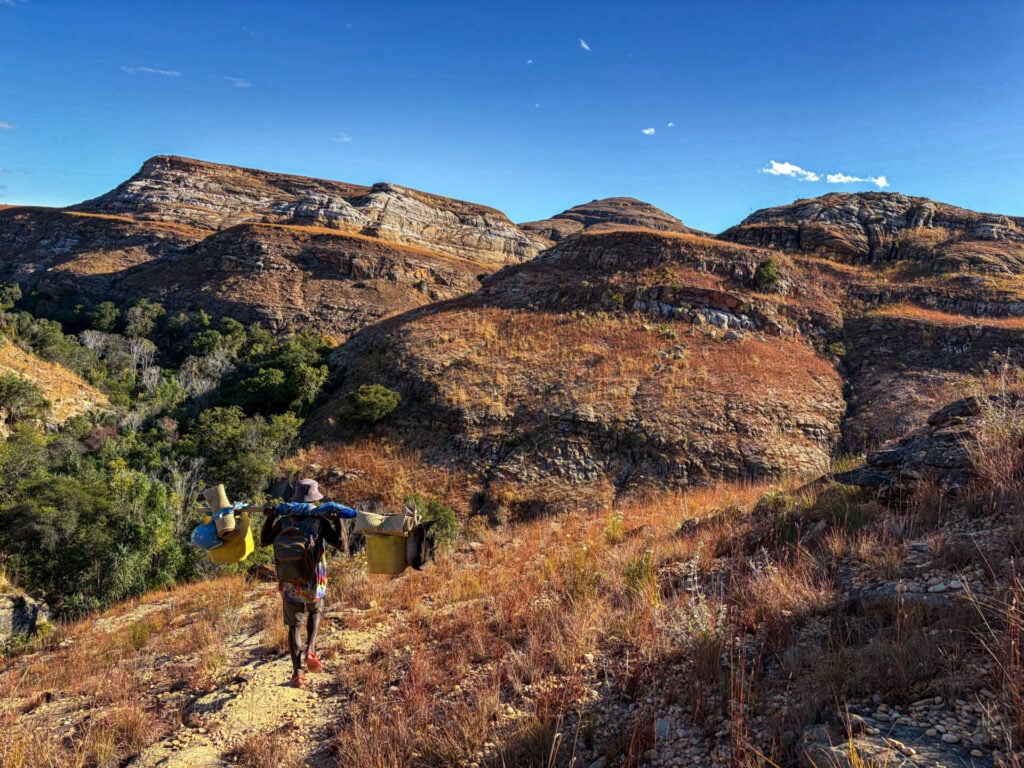 Une personne se lance dans un voyage mystère, marchant le long d'un sentier aride et herbeux à flanc de colline à Madagascar, chargée de conteneurs et de sacs, avec en arrière-plan des collines rocheuses et une végétation clairsemée, sous un ciel bleu limpide. - Karavaniers