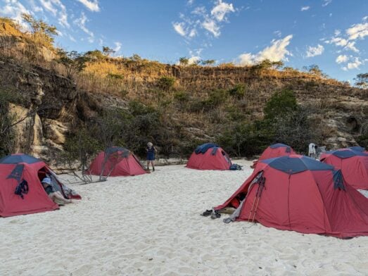 Plusieurs tentes rouges sont dressées sur un sol sablonneux, près d'un terrain rocheux recouvert de végétation sèche, évoquant un voyage mystérieux à Madagascar. Des bâtons de randonnée et des sacs à dos sont posés près des tentes, tandis qu'une personne se tient debout à l'arrière-plan, sous un ciel partiellement nuageux. - Karavaniers