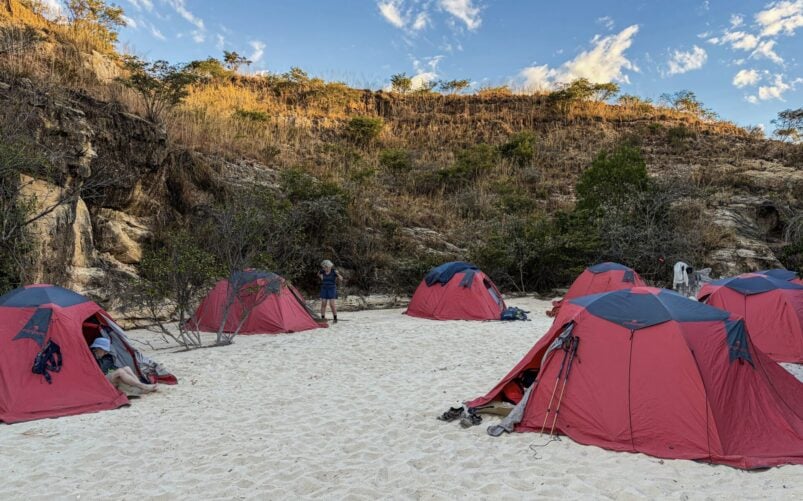 Plusieurs tentes rouges sont dressées sur un sol sablonneux, près d'un terrain rocheux recouvert de végétation sèche, évoquant un voyage mystérieux à Madagascar. Des bâtons de randonnée et des sacs à dos sont posés près des tentes, tandis qu'une personne se tient debout à l'arrière-plan, sous un ciel partiellement nuageux. - Karavaniers