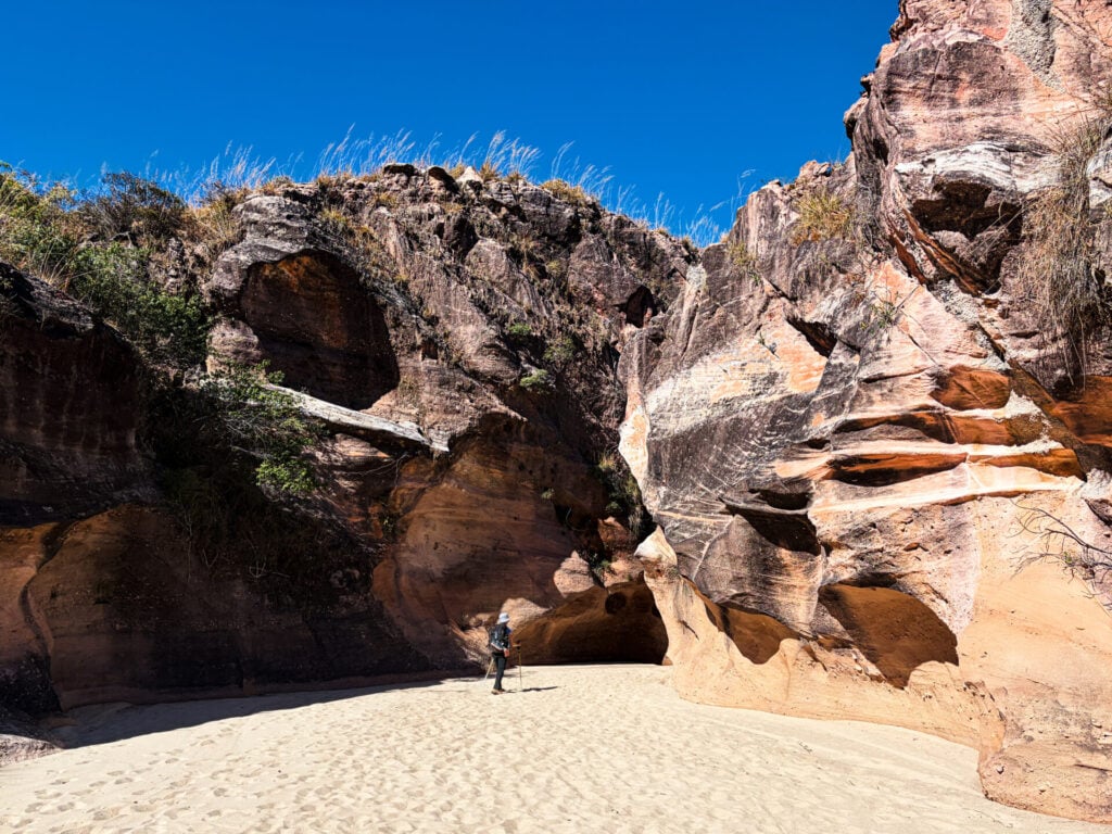 Une personne se tient debout sur un sol sablonneux, entre de hautes formations rocheuses escarpées aux motifs stratifiés, sous un ciel bleu limpide — une image qui incarne l'esprit d'un voyage mystérieux à Madagascar. Une végétation clairsemée pousse au sommet des rochers. - Karavaniers