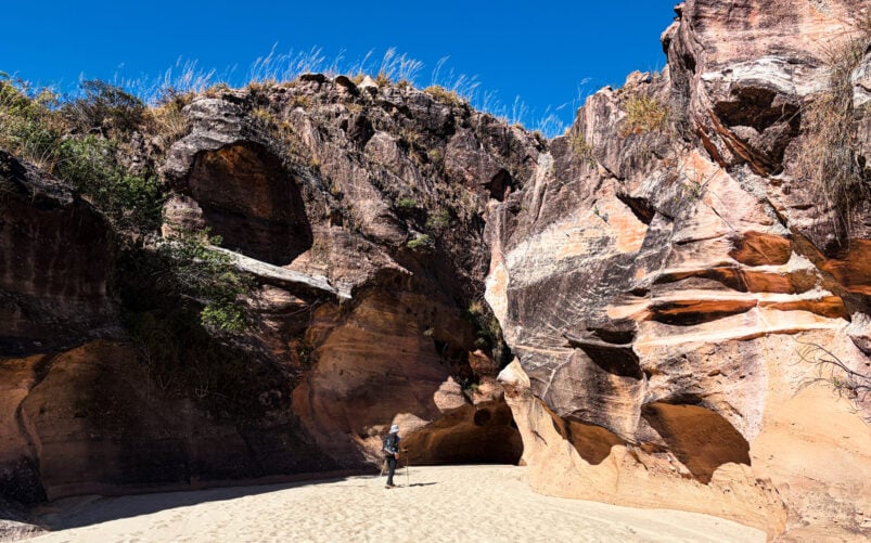 Une personne se tient debout sur un sol sablonneux, entre de hautes formations rocheuses escarpées aux motifs stratifiés, sous un ciel bleu limpide — une image qui incarne l'esprit d'un voyage mystérieux à Madagascar. Une végétation clairsemée pousse au sommet des rochers. - Karavaniers