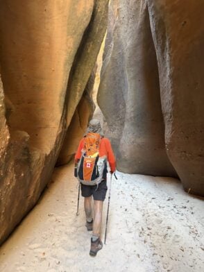 Une personne vêtue d'un t-shirt orange, d'un short de randonnée et portant un sac à dos orné du drapeau canadien se lance dans un voyage mystère à travers un canyon étroit et sablonneux aux hautes parois rocheuses. Des bâtons de randonnée la guident sur ce sentier plein d'aventures. - Karavaniers