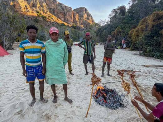 Plusieurs personnes se tiennent sur un sol sablonneux près d'un feu de camp où de la viande rôtit sur des brochettes : une scène tirée d'un « voyage mystère » à Madagascar. Des falaises rocheuses et des arbres se dressent en arrière-plan, tandis qu'une personne est accroupie près du feu et que d'autres se rassemblent à proximité. - Karavaniers