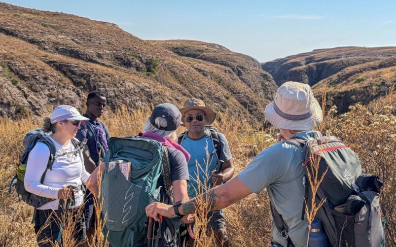 Cinq randonneurs, coiffés de chapeaux et munis de sacs à dos, se tiennent dans un paysage aride et herbeux, parsemé de collines ondulantes et d’un canyon, qui rappelle un « Voyage mystère » à Madagascar. Ils semblent discuter ou planifier la suite de leur parcours sous un ciel bleu limpide. - Karavaniers