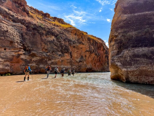 Cinq randonneurs, équipés de sacs à dos et de bâtons de randonnée, se lancent dans un voyage mystère, marchant au fond d'une rivière peu profonde entre les hautes parois rocheuses d'un canyon, sous le ciel d'un bleu éclatant de Madagascar. - Karavaniers
