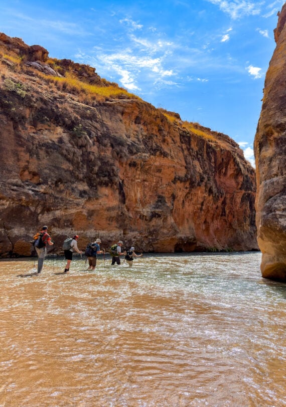 Cinq randonneurs, équipés de sacs à dos et de bâtons de randonnée, se lancent dans un voyage mystère, marchant au fond d'une rivière peu profonde entre les hautes parois rocheuses d'un canyon, sous le ciel d'un bleu éclatant de Madagascar. - Karavaniers