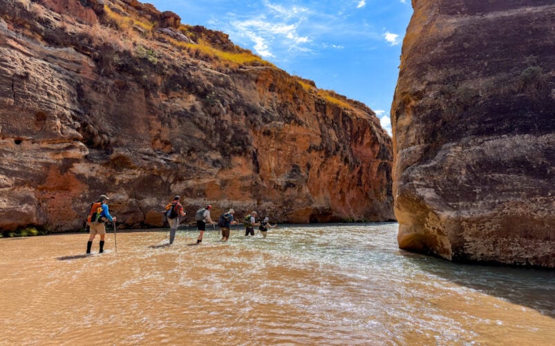 Cinq randonneurs, équipés de sacs à dos et de bâtons de randonnée, se lancent dans un voyage mystère, marchant au fond d'une rivière peu profonde entre les hautes parois rocheuses d'un canyon, sous le ciel d'un bleu éclatant de Madagascar. - Karavaniers