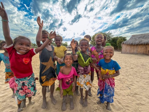 Un groupe d'enfants souriants, pieds nus sur le sable, lèvent les bras de joie lors d'un « voyage mystère » à Madagascar, avec en arrière-plan une hutte au toit de chaume et une végétation luxuriante, sous un ciel partiellement nuageux. - Karavaniers