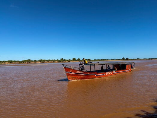 Un bateau de passagers rouge et noir portant l'inscription « BOLIVIA » entame un voyage mystère sur un large fleuve aux eaux brunes, sous un ciel bleu limpide, tandis qu'une végétation basse et verte se profile au loin le long des rives. - Karavaniers