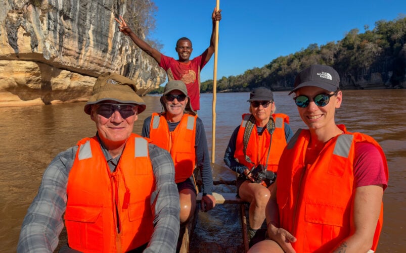 Cinq personnes vêtues de gilets de sauvetage orange vif sont assises dans un petit bateau sur une rivière, près de falaises rocheuses, sous un ciel bleu limpide : un voyage mystère à travers la beauté sauvage de Madagascar, avec une personne debout à l'arrière qui guide le groupe. - Karavaniers