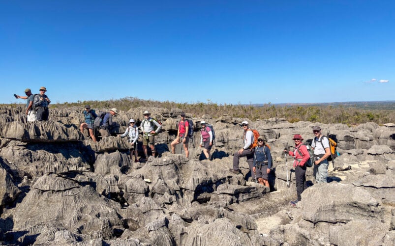 Un groupe de personnes, coiffées de chapeaux et munies de sacs à dos, se lance dans un voyage mystère, marchant et avançant sur un terrain accidenté et rocailleux sous un ciel bleu limpide à Madagascar, avec de la végétation basse visible en arrière-plan. - Karavaniers