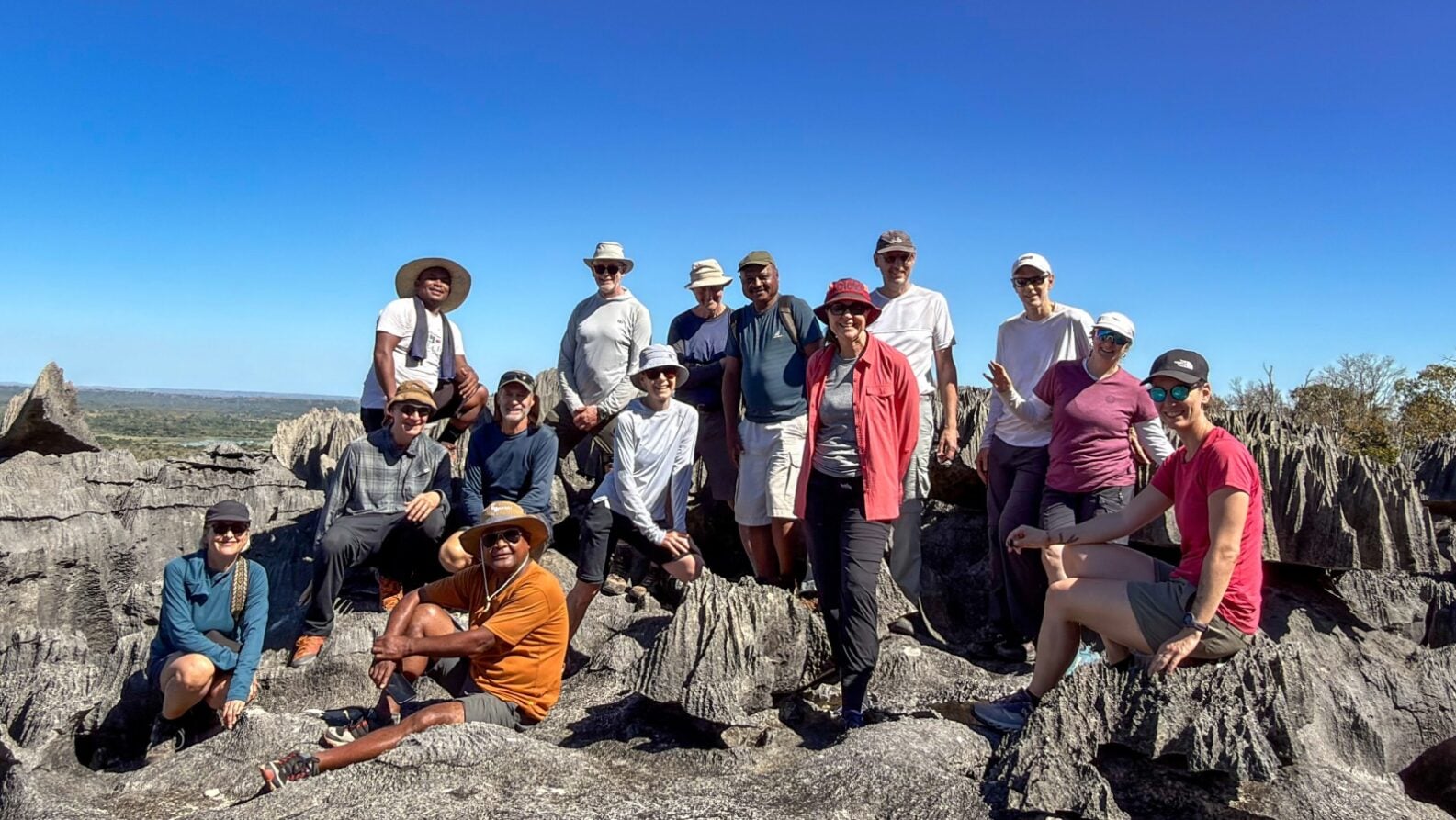 Un groupe de quinze personnes pose ensemble sur des formations rocheuses escarpées, sous un ciel bleu limpide, coiffées de chapeaux et vêtues de tenues décontractées de plein air — incarnant ainsi l'esprit d'un « voyage mystère » à Madagascar, au milieu de paysages rocheux et d'une végétation clairsemée. - Karavaniers