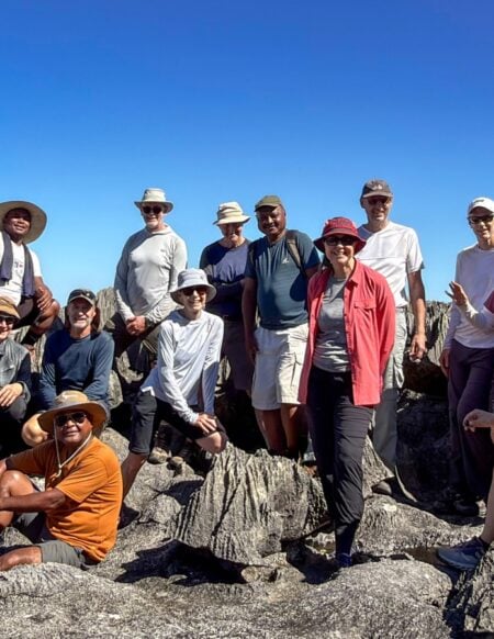 Un groupe de quinze personnes pose ensemble sur des formations rocheuses escarpées, sous un ciel bleu limpide, coiffées de chapeaux et vêtues de tenues décontractées de plein air — incarnant ainsi l'esprit d'un « voyage mystère » à Madagascar, au milieu de paysages rocheux et d'une végétation clairsemée. - Karavaniers