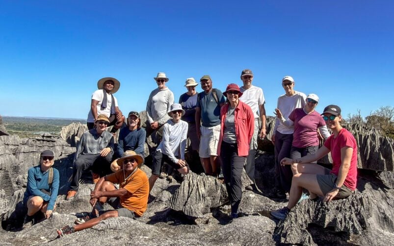 Un groupe de quinze personnes pose ensemble sur des formations rocheuses escarpées, sous un ciel bleu limpide, coiffées de chapeaux et vêtues de tenues décontractées de plein air — incarnant ainsi l'esprit d'un « voyage mystère » à Madagascar, au milieu de paysages rocheux et d'une végétation clairsemée. - Karavaniers