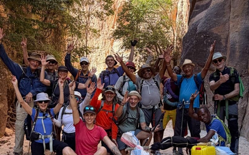 Un groupe de randonneurs prend la pose et sourit pour une photo dans un canyon sablonneux, entouré de hautes parois rocheuses et d'arbres, incarnant ainsi l'esprit d'une aventure « Voyage Mystère » à Madagascar. La plupart se tiennent debout, les mains levées, tandis que quelques-uns sont assis au premier plan, avec leurs sacs à dos et leur équipement de randonnée. - Karavaniers