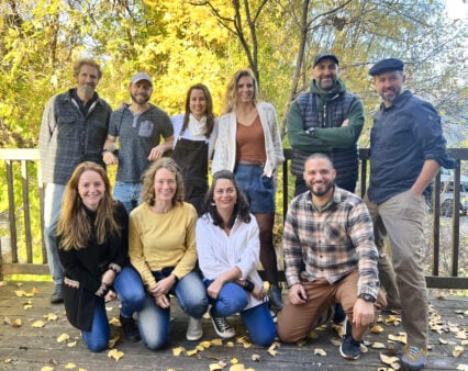Un groupe de dix adultes pose ensemble en plein air sur une terrasse en bois entourée d'arbres aux feuilles d'automne jaunes. Trois personnes sont agenouillées devant, tandis que les autres sont debout derrière elles, toutes face à l'appareil photo et souriantes. - Karavaniers