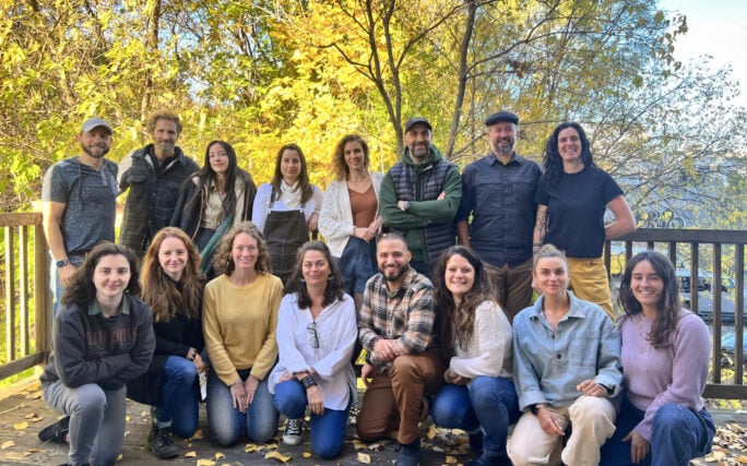 Un groupe de seize personnes pose ensemble en plein air sur une terrasse en bois entourée d'arbres au feuillage automnal. Certaines sont debout, d'autres sont agenouillées ou assises par terre, toutes font face à l'appareil photo et sourient. - Karavaniers