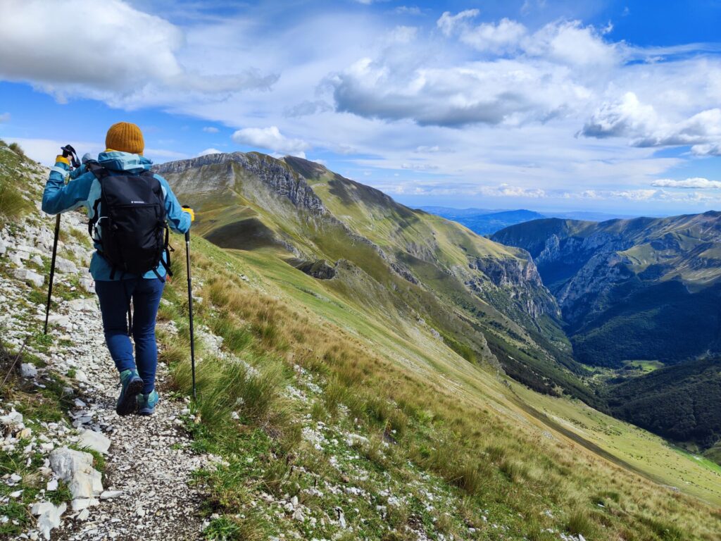 Une personne vêtue d'une veste bleue et d'un bonnet orange fait une randonnée sur un sentier de montagne rocailleux, équipée de bâtons de randonnée. En arrière-plan, on aperçoit des montagnes, des pentes herbeuses et un ciel partiellement nuageux. - Karavaniers