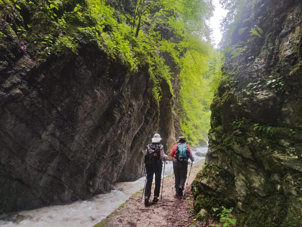 Deux randonneurs, équipés de sacs à dos, marchent sur un sentier étroit et rocailleux qui longe un ruisseau, entourés de falaises escarpées et moussues ainsi que d'une végétation dense et verdoyante, sous un ciel nuageux. - Karavaniers