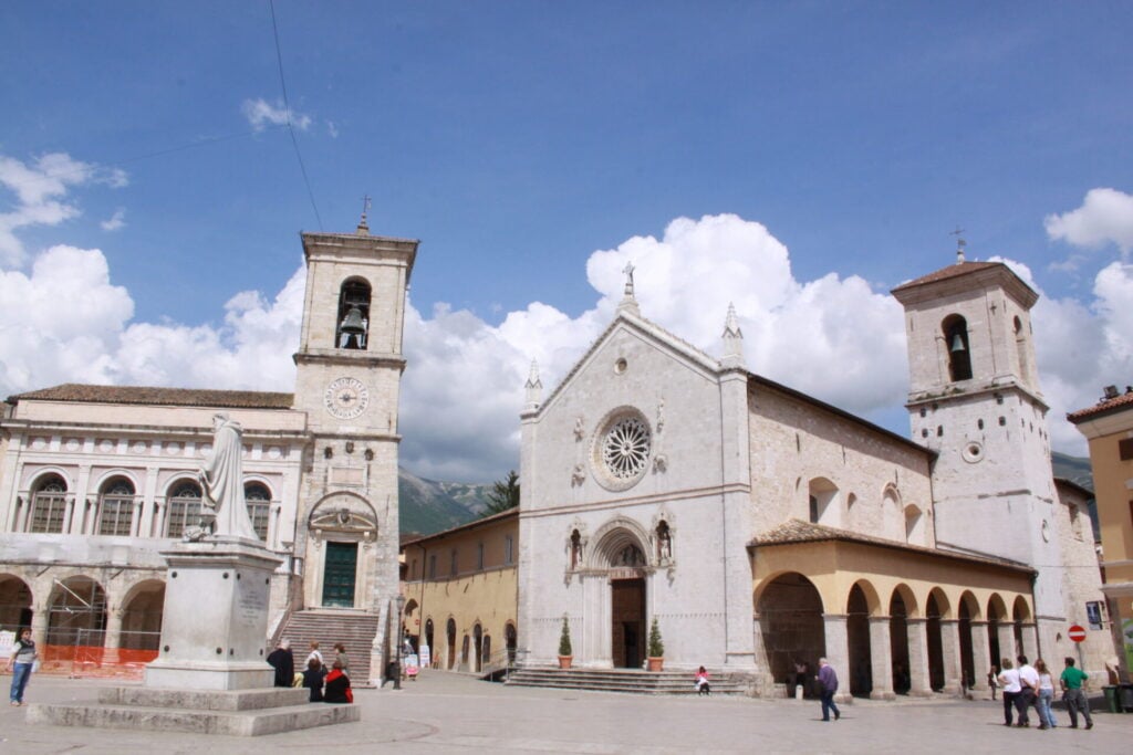 Une cathédrale en pierre, dotée d'une rosace et de deux clochers, se dresse sur la place de la ville. Des gens se promènent dans les environs, et une statue posée sur un socle est visible au premier plan. Le ciel est bleu, parsemé de quelques nuages. - Karavaniers