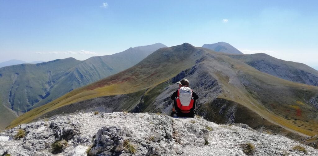 Une personne portant un sac à dos rouge est assise sur un terrain rocheux, de dos, et contemple une chaîne de montagnes vertes et grises sous un ciel bleu limpide. - Karavaniers