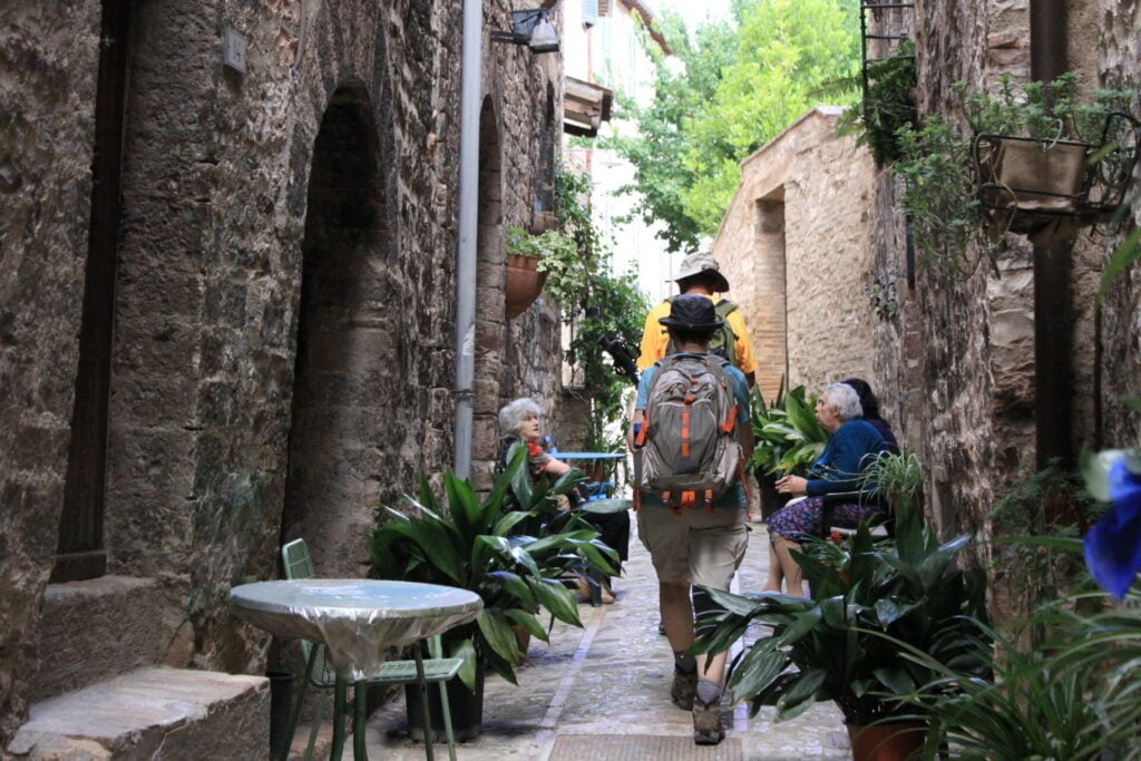Deux personnes, munies de sacs à dos, descendent une ruelle étroite pavée, bordée de plantes en pot. Trois personnes d'un certain âge sont assises à une petite table sur la gauche et discutent. On aperçoit de la verdure et des rayons de soleil au bout de la ruelle. - Karavaniers