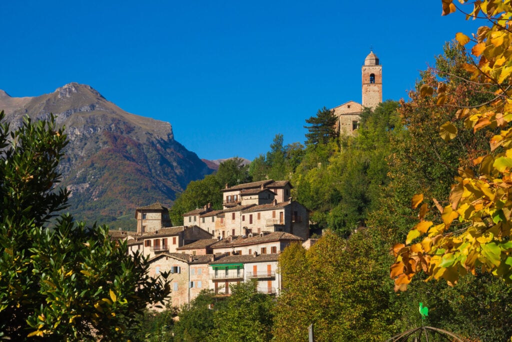 Un village à flanc de colline, avec ses maisons en pierre et sa tour horloge, est entouré d'arbres verdoyants et de feuillage automnal, avec une montagne et un ciel bleu limpide en arrière-plan. - Karavaniers
